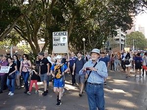 March for Science protesters in Hyde Park, Sydney. An older White man is in the foreground. Behind him someone holds a sign that reads 
