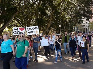 March for Science protesters walk through Hyde Park, Sydney. A White, bearded man wears a bright green t-shirt in the front. Behind him one person holds a sign 